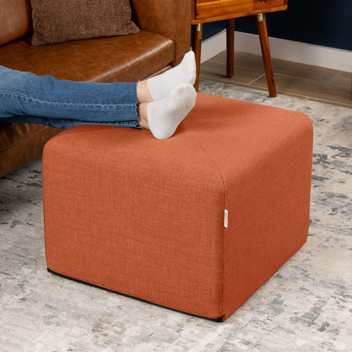 Jaxx Monroe 24in foam ottoman in canyon orange performance fabric, used as a footrest in a living room setting