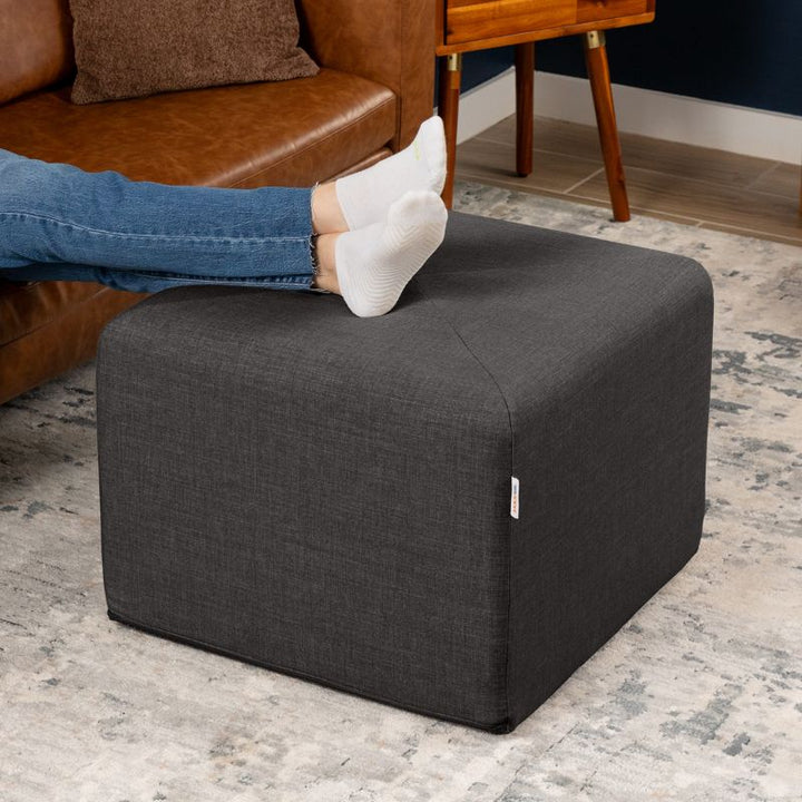 Jaxx Monroe 24in foam ottoman in charcoal performance fabric, used as a footrest in a living room setting