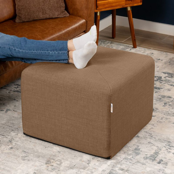 Jaxx Monroe 24in foam ottoman in earth brown performance fabric, used as a footrest in a living room setting