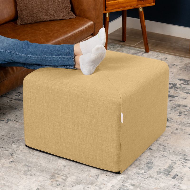 Jaxx Monroe 24in foam ottoman in wheat gold performance fabric, used as a footrest in a living room setting