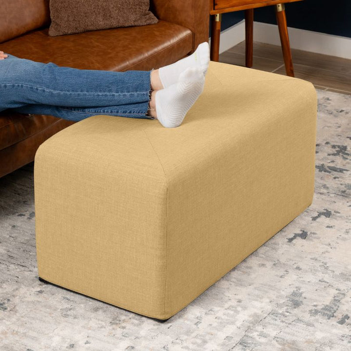 Jaxx Monroe 36in rectangular foam ottoman in wheat gold performance fabric, used as a footrest in a living room setting