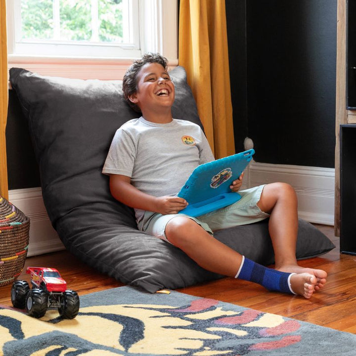 Laughing boy with a tablet leaning back on a Jaxx Pillow Saxx 4 bean bag floor pillow in Black, propped against the wall as a cozy kids floor pillow