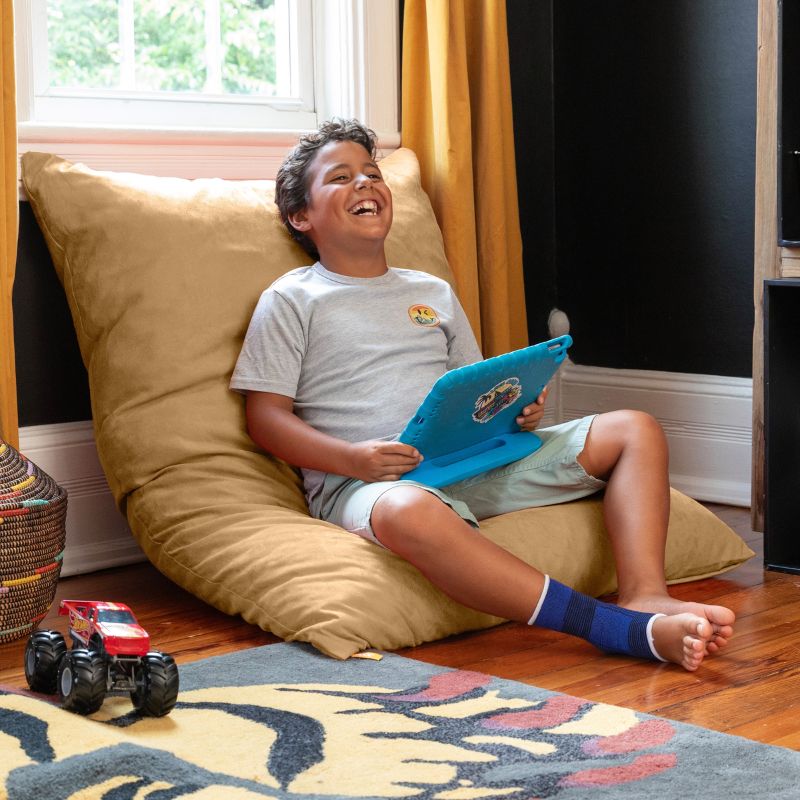 Boy relaxing with a tablet on a Jaxx Pillow Saxx 4 bean bag floor pillow in Camel, set upright against the wall in a playroom