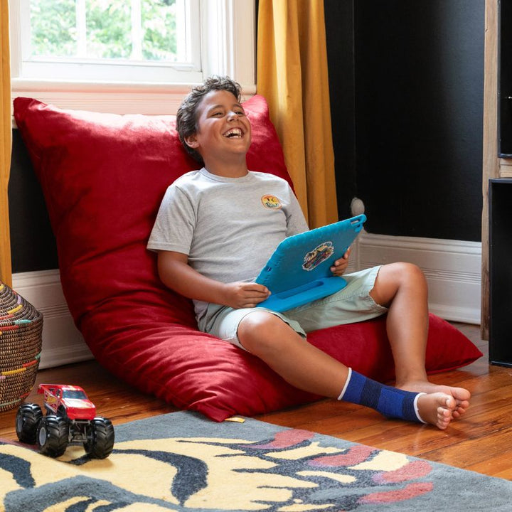 Smiling boy leaning back on a Jaxx Pillow Saxx 4 bean bag floor pillow in Cherry, a bright kids floor cushion for screen time and downtime
