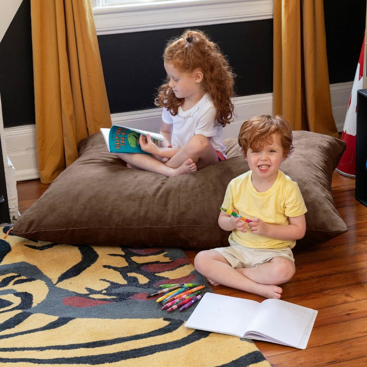 Two kids reading and colouring on and beside a Chocolate Jaxx Pillow Saxx 4 kids floor pillow in a cozy bedroom corner