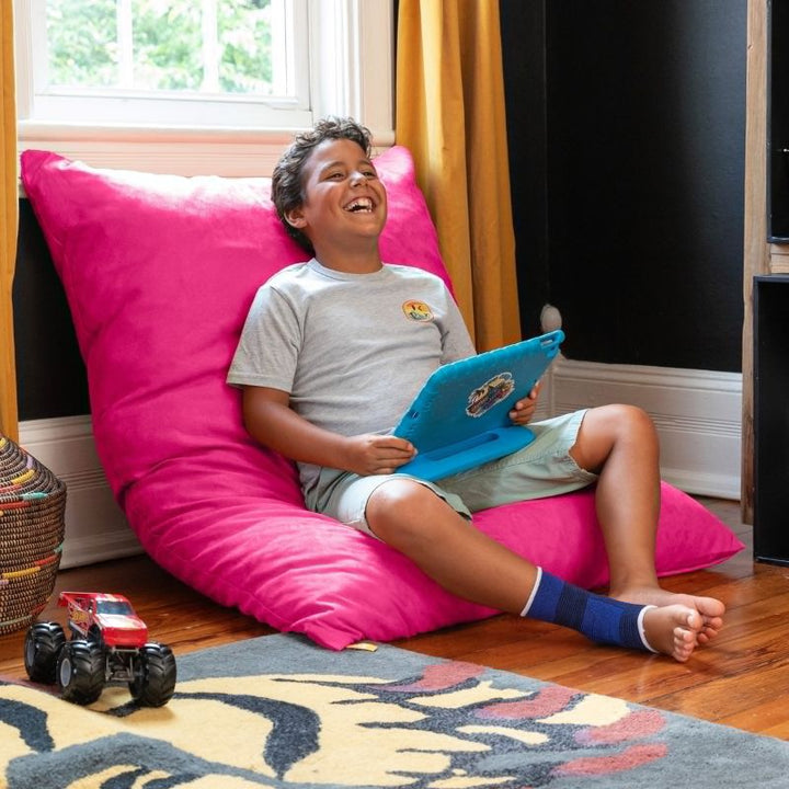 Boy laughing with a tablet on a Jaxx Pillow Saxx 4 bean bag floor pillow in Fuchsia, propped upright as a colourful kids floor cushion