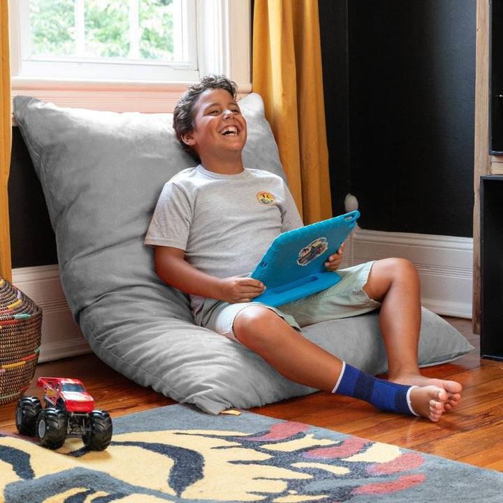 Smiling boy with a tablet leaning on a Jaxx Pillow Saxx 4 bean bag floor pillow in Platinum, a soft neutral kids floor pillow for homes and classrooms