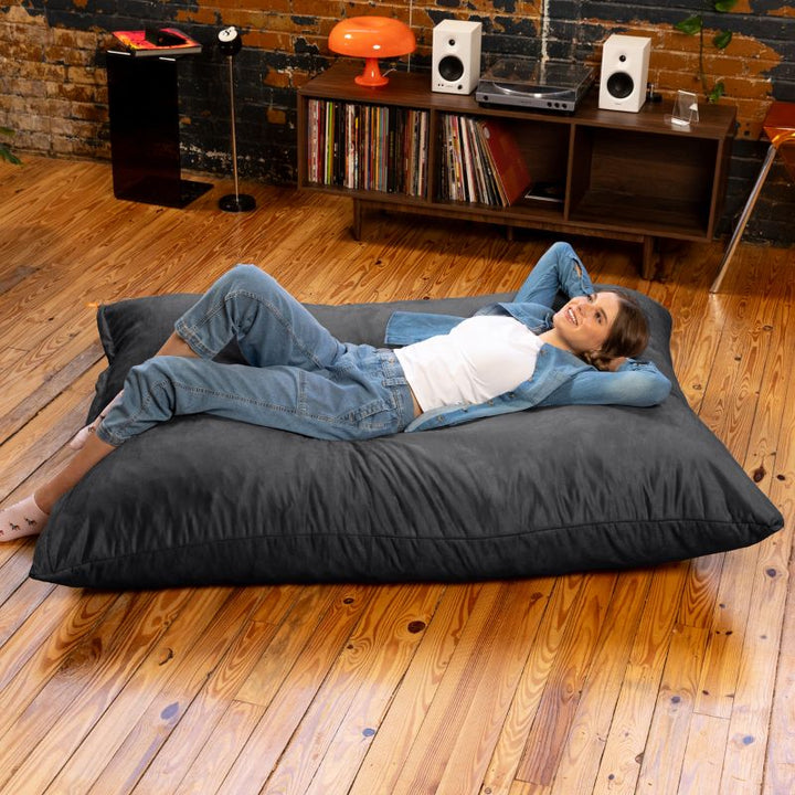 Woman lying back with her hands behind her head on a Jaxx Pillow Saxx 5 giant bean bag floor pillow in Black, stretched out in a record-filled living room