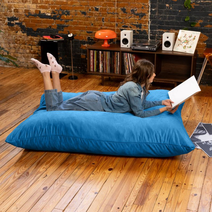 Woman reading on a Blueberry Jaxx Pillow Saxx 5 bean bag floor pillow, an oversized floor cushion in a relaxed loft-style space