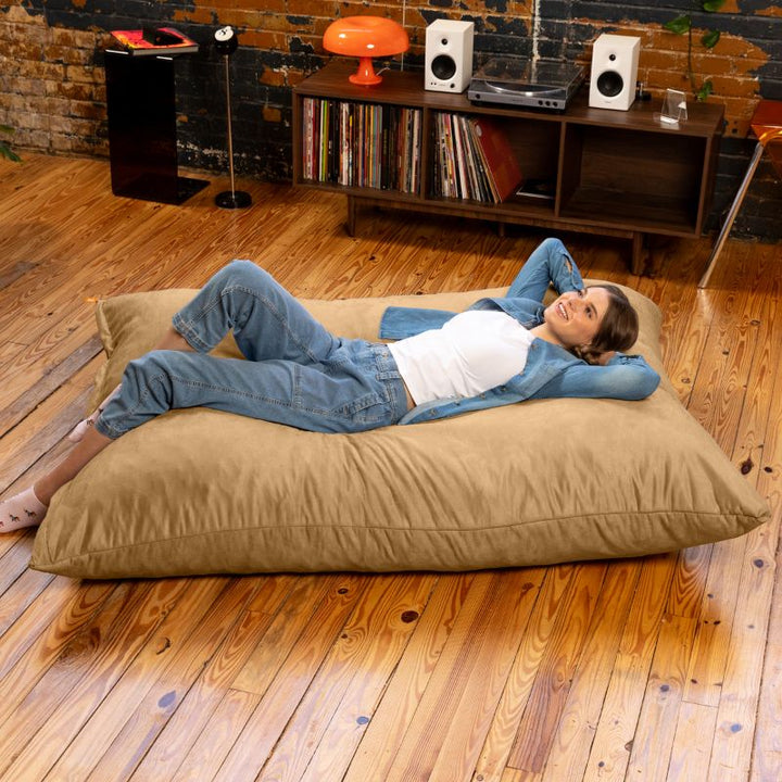 Adult unwinding flat on a Jaxx Pillow Saxx 5 giant bean bag floor pillow in Camel, a warm neutral crash pillow for family rooms