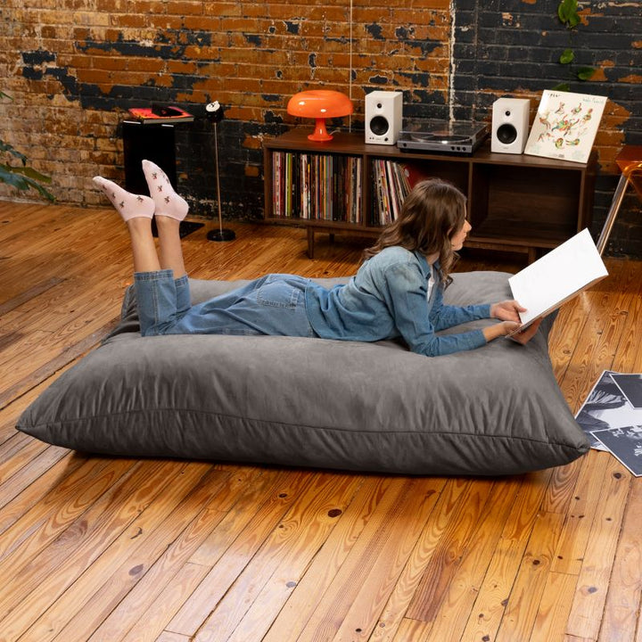 Reader lying on a Charcoal Jaxx Pillow Saxx 5 bean bag floor pillow on a hardwood floor, a giant floor cushion for rec rooms