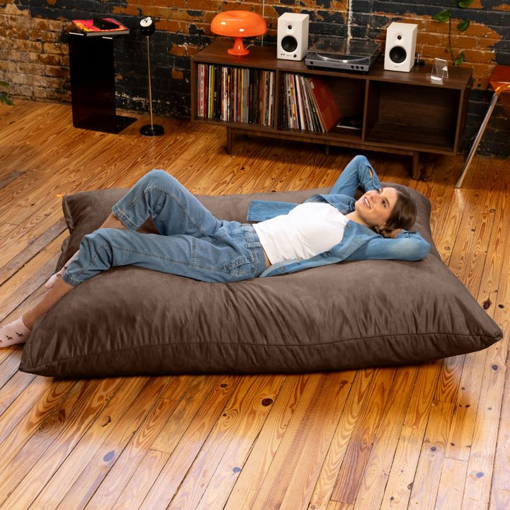 Woman relaxing on her back on a Jaxx Pillow Saxx 5 giant bean bag floor pillow in Chocolate, room to fully stretch out on an oversized floor cushion