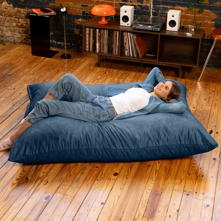 Adult resting on her back on a Jaxx Pillow Saxx 5 giant bean bag floor pillow in Navy, a deep-toned crash pillow for lounging