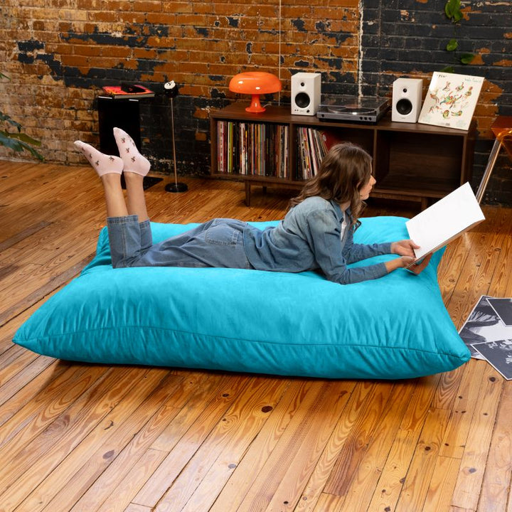 Reader relaxing on a Teal Jaxx Pillow Saxx 5 bean bag floor pillow, a vibrant giant crash pillow for rec rooms and calm corners