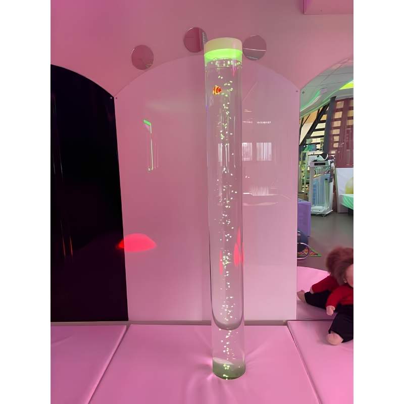 A floor-standing bubble tube with a colour-changing light base and a clownfish figure inside the water column, set up in a soft play sensory room with small round wall mirrors visible in the background
