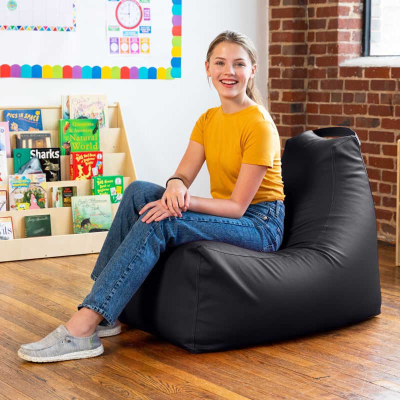 Student sitting on the Jaxx Pezzi classroom bean bag chair in black vinyl beside a bookshelf in a classroom reading nook