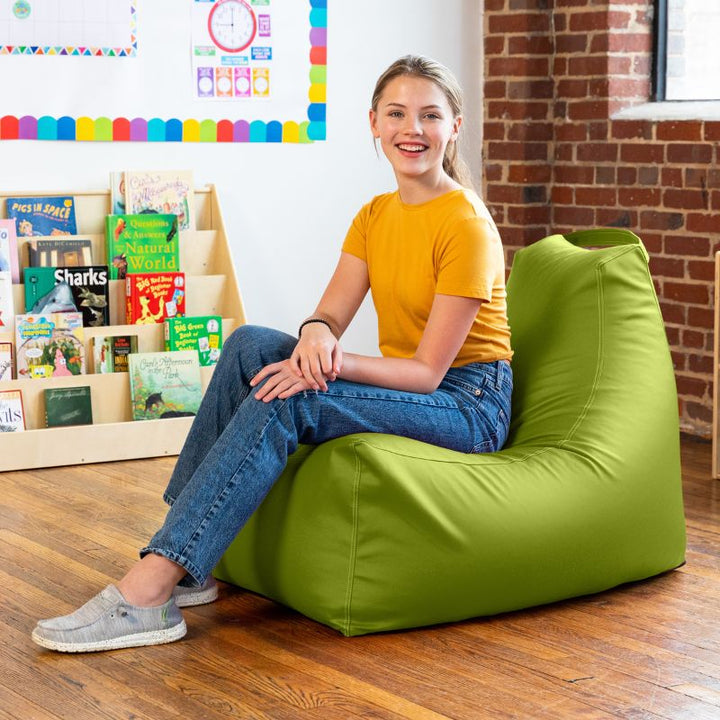 Student relaxing on the Jaxx Pezzi classroom bean bag chair in green vinyl in a bright school library corner