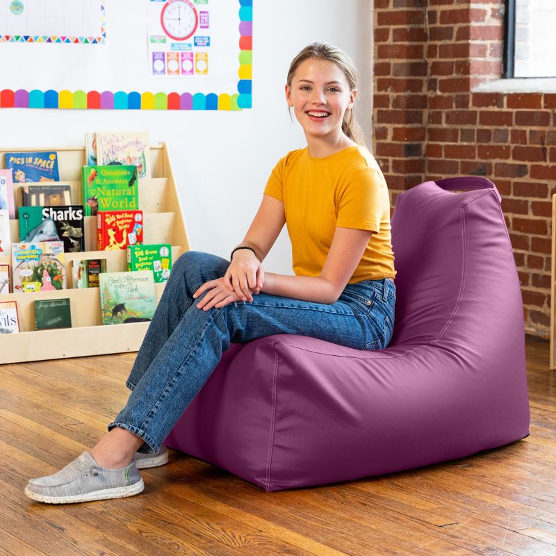Student seated on the Jaxx Pezzi classroom bean bag chair in purple vinyl beside classroom bookshelves