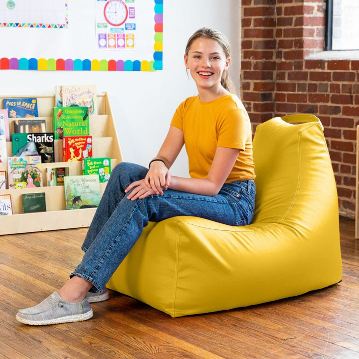Student relaxing on the Jaxx Pezzi classroom bean bag chair in yellow vinyl beside classroom bookshelves
