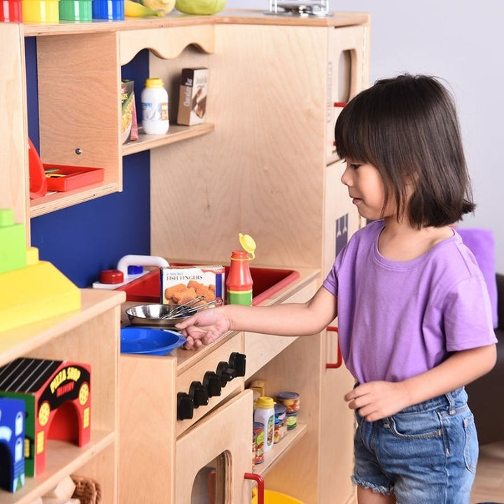 Child plating pretend food at the Constructive Playthings Five-In-One Kitchen, showing the dramatic play kitchen in use for role-play and fine motor development in a classroom setting