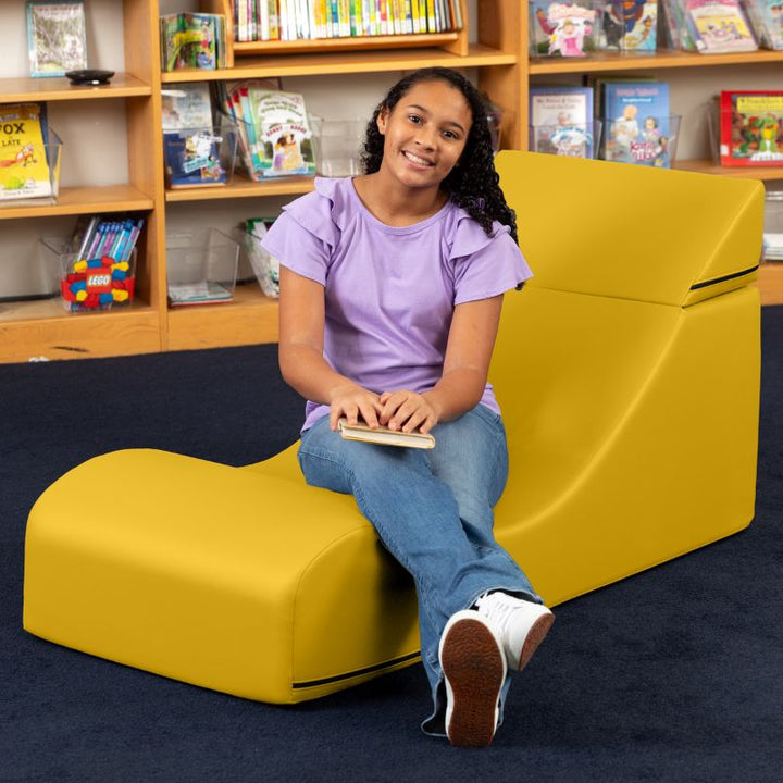 A student seated in the Jaxx Flexx convertible foam chair in yellow vinyl in a school library setting