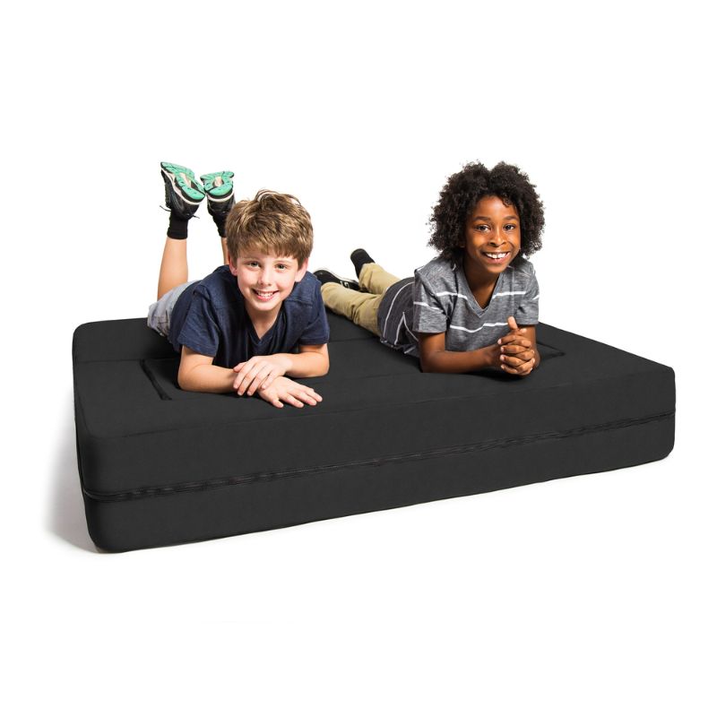 Two kids relaxing on the Jaxx Zipline flip sofa in black, shown flattened into its bed configuration