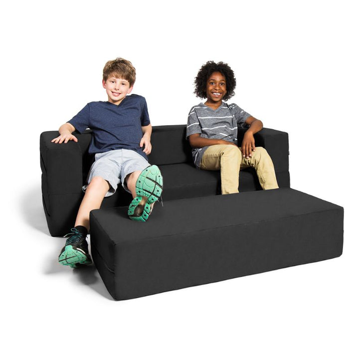 Two kids sitting on the Jaxx Zipline convertible kids sofa and ottoman in black, showing the seat depth in its loveseat configuration