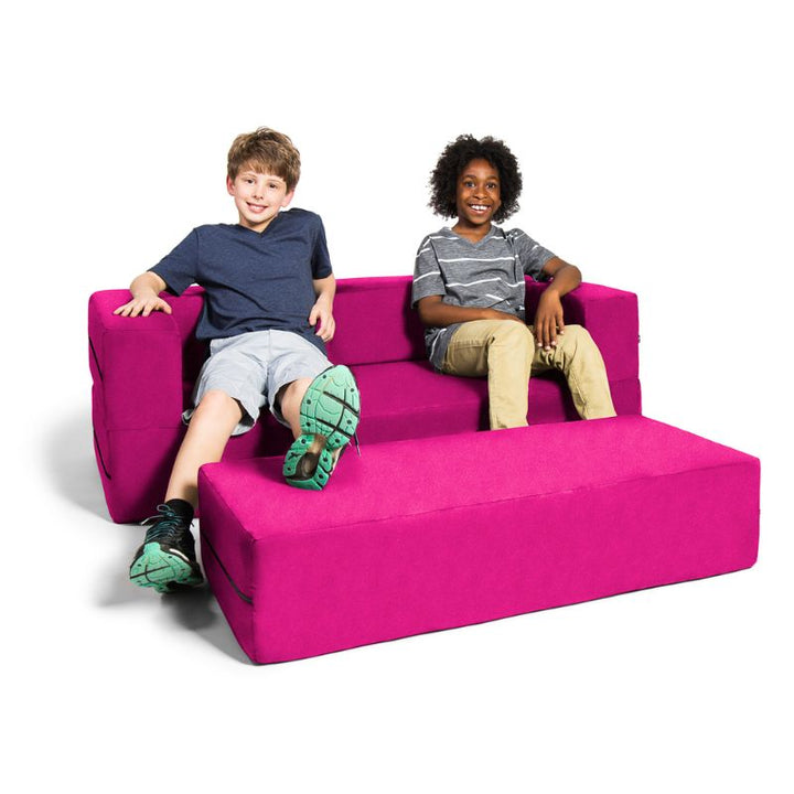 Two kids sitting on the Jaxx Zipline convertible kids sofa and ottoman in pink, showing the flip sofa at true kid scale