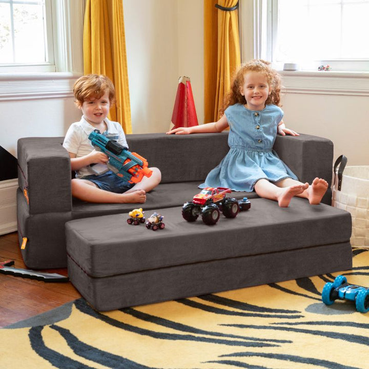 Two kids sitting on the Jaxx Zipline flip sofa in charcoal, shown in a playroom with toys on the ottoman