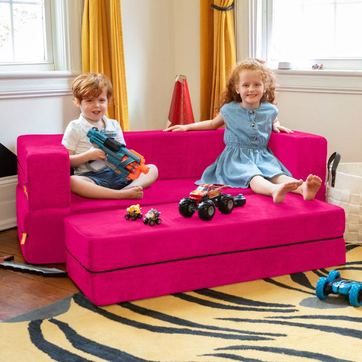 Two kids sitting on the Jaxx Zipline convertible kids sofa and ottoman in pink, playing in a bedroom setting