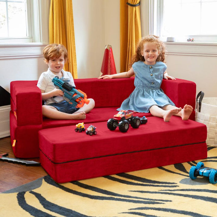 Two kids sitting on the Jaxx Zipline convertible kids sofa in red, with toys spread across the ottoman