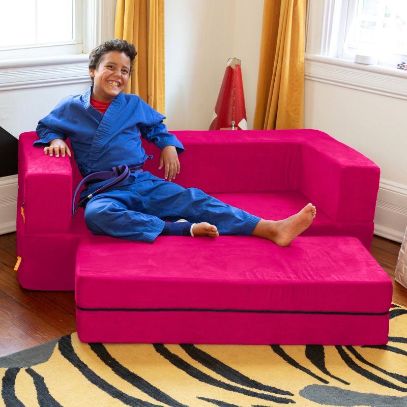 A boy sitting on the Jaxx Zipline convertible kids sofa and ottoman in pink, styled in a bedroom lifestyle shot