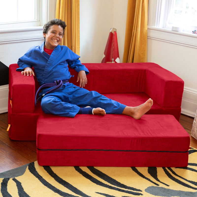A boy sitting on the Jaxx Zipline convertible kids sofa and ottoman in red, styled in a bedroom