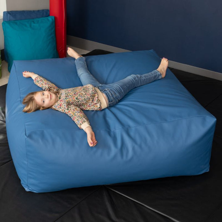 A young girl relaxes on the blue Jaxx Crash Mat 48in in a school sensory room, showing the crash mat's full-body landing surface