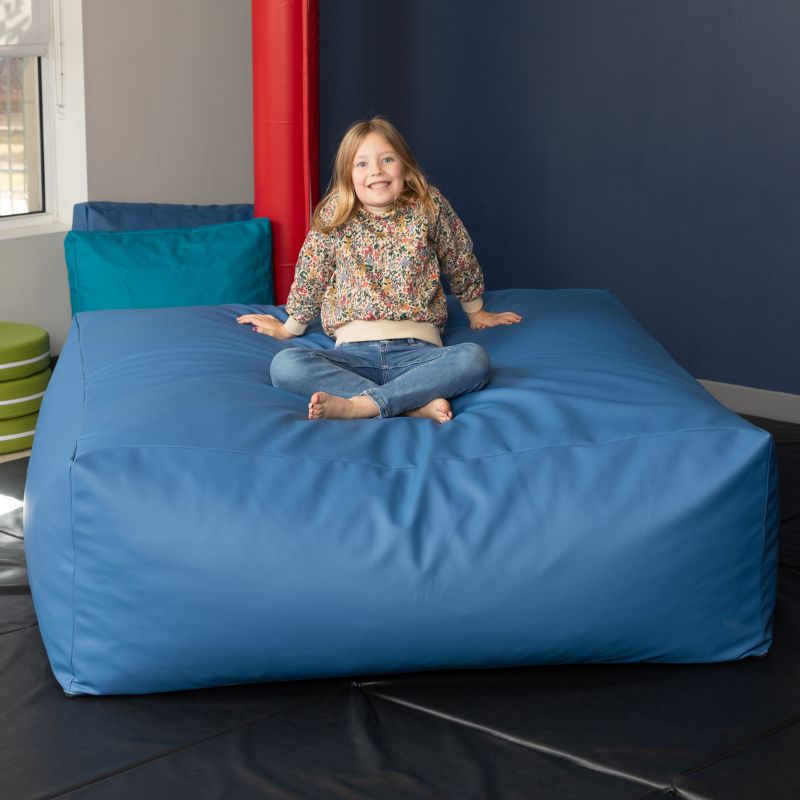 A young girl sits cross-legged on the blue Jaxx Crash Mat 48in, demonstrating the crash mat as a cozy floor cushion for calm-down time