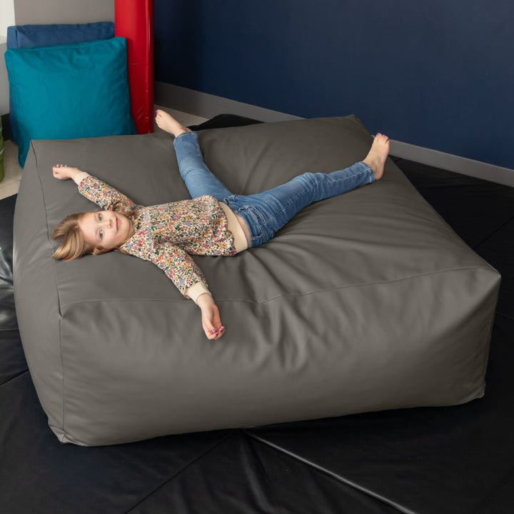 A young girl relaxes on the charcoal Jaxx Crash Mat 48in in a school sensory room, showing the crash mat's full-body landing surface