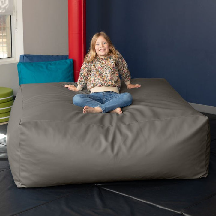 A young girl sits cross-legged on the charcoal Jaxx Crash Mat 48in, demonstrating the crash mat as a cozy floor cushion for calm-down time
