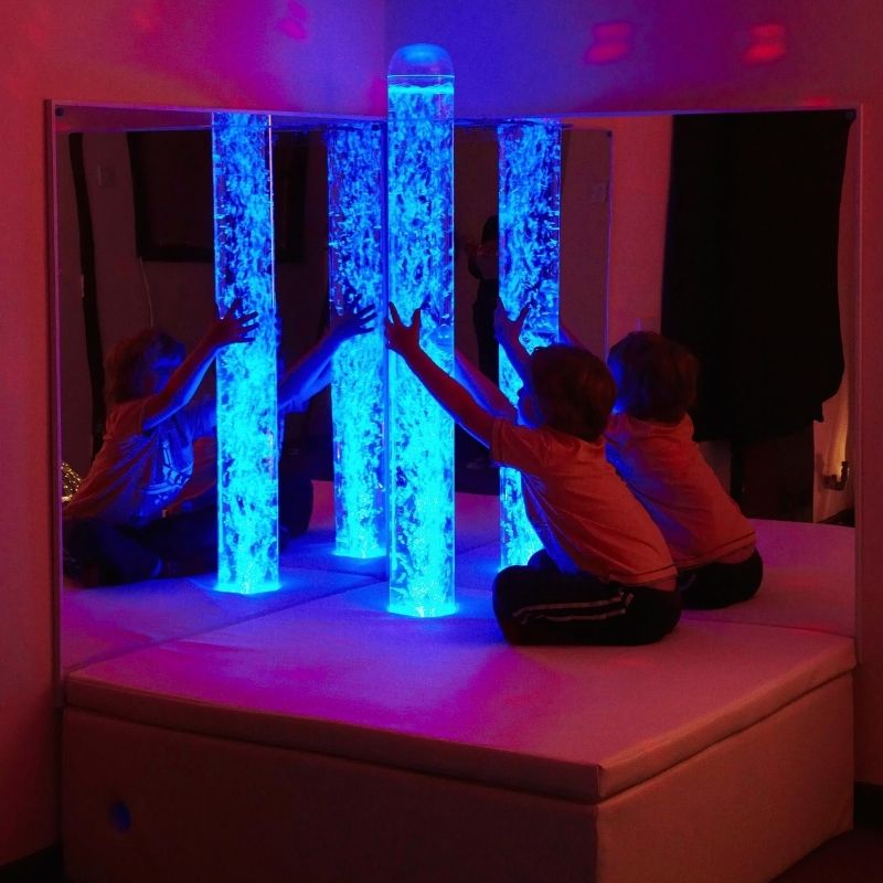 Child actively engaged with the Experia Bubble Tube Mirrors corner installation in blue light, showing the quadrupled visual experience of the shatter-resistant acrylic mirror pair