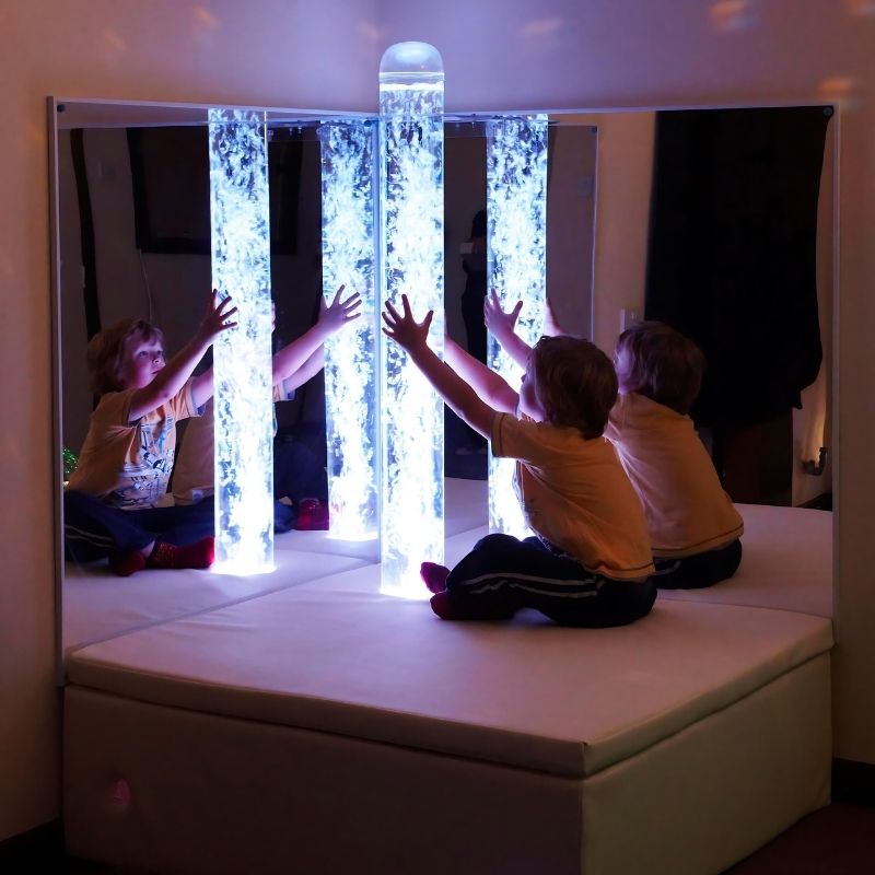 Child playing beside the Experia Bubble Tube Mirrors in cool white light, showing the immersive corner sensory environment created by the acrylic mirror pair