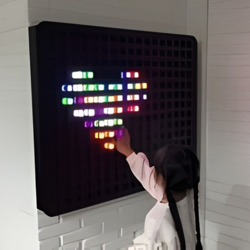 Back view of a child placing coloured pegs on the Experia PixelPop Interactive Light Panel, showing the wall-mounted installation and glowing peg design in progress