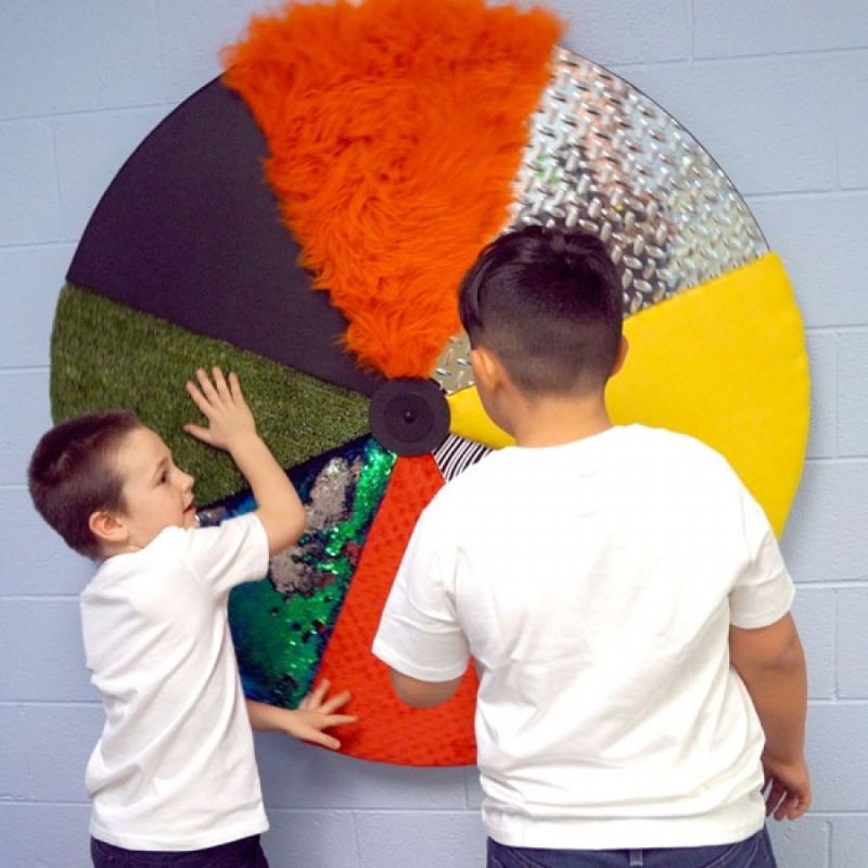 Two children exploring the Experia Sensory Texture Wheel together, touching the fur, artificial turf, colour-changing sequin and diamond plate panels of the 46" wall-mounted tactile wheel
