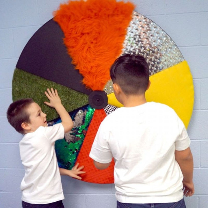 Two children exploring the Experia Sensory Texture Wheel together, touching the fur, artificial turf, colour-changing sequin and diamond plate panels of the 46" wall-mounted tactile wheel