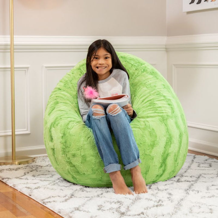 A child sitting on the Jaxx Cocoon Fur faux fur bean bag chair in Green, styled in a bedroom as a cozy reading spot
