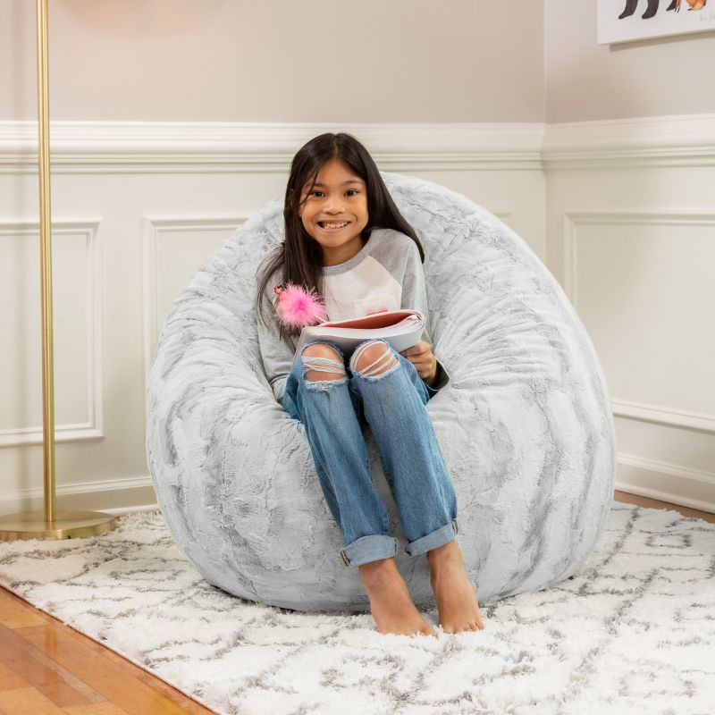 A child sitting on the Jaxx Cocoon Fur faux fur bean bag chair in Grey, styled in a bedroom as a cozy reading spot
