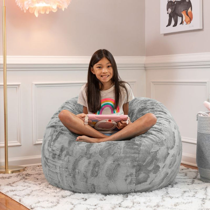Girl relaxing on the Jaxx Saxx faux fur bean bag chair in Grey in a bedroom setting, a soft tactile sensory seat for kids