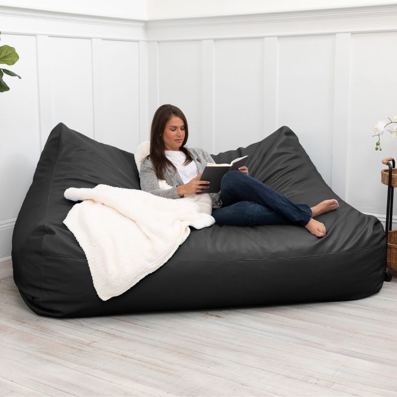 A woman reading on the Jaxx Lozia faux leather loveseat in black, a two-seat classroom soft seating option in a home setting