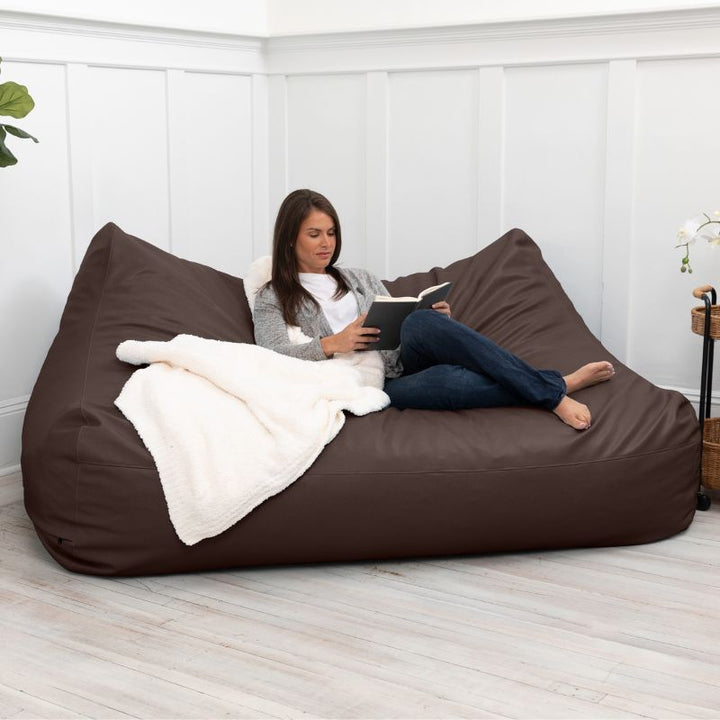 A woman reading on the Jaxx Lozia faux leather loveseat in chocolate, easy-clean waiting room seating