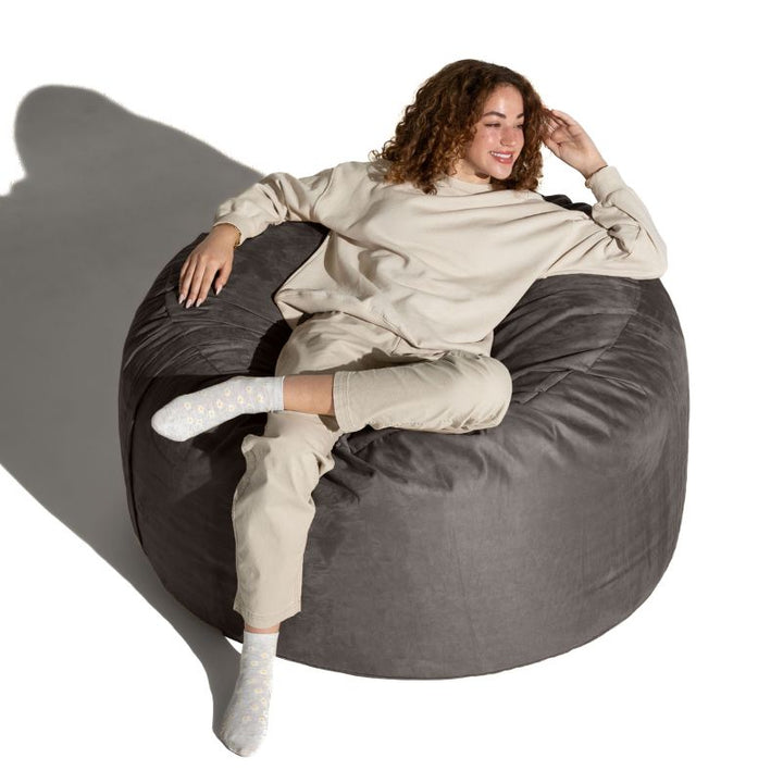 Woman stretched out on a Charcoal microsuede Jaxx Saxx 4ft foam bean bag chair, showing how the foam fill supports the whole body