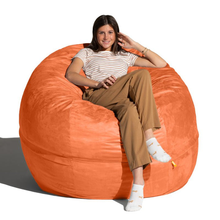 Smiling young woman sitting in a Mandarin microsuede Jaxx Saxx 5ft foam bean bag chair, a bright large bean bag chair for rec rooms