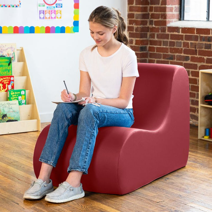Student sitting in a claret dark red Jaxx Midtown foam chair while writing, a wipeable classroom soft seating chair for kids and adults